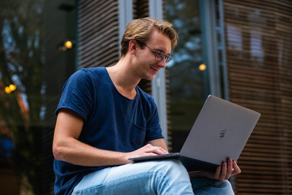 A young student smiling while using a budgeting spreadsheet on their laptop in a sunlit dorm room.
