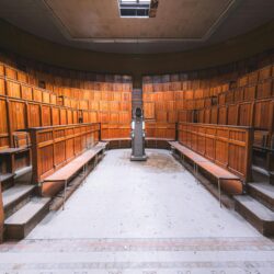 A focused young student sitting in a tiered lecture hall, writing in a notebook with their laptop open.
