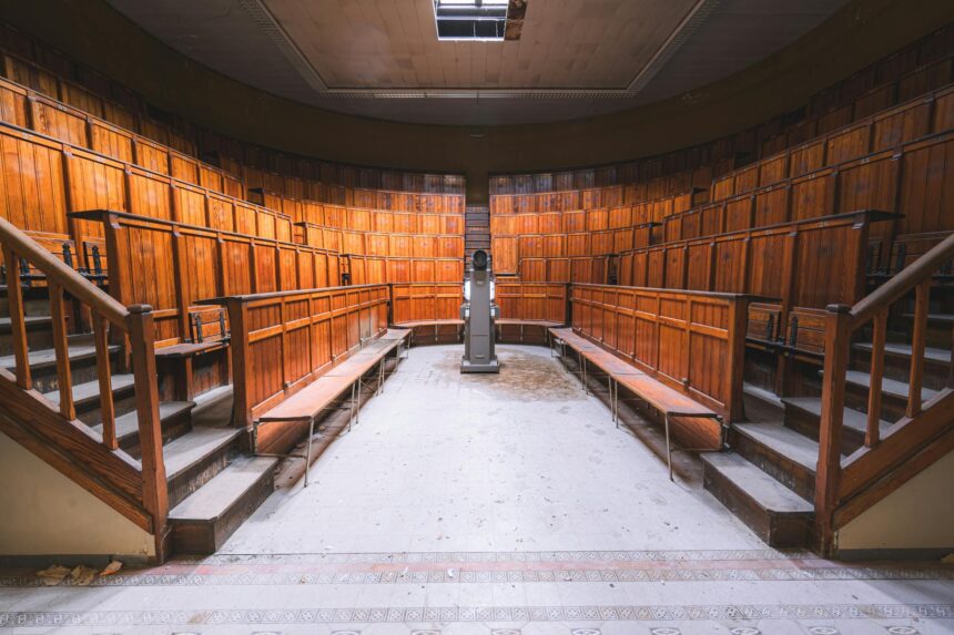 A focused young student sitting in a tiered lecture hall, writing in a notebook with their laptop open.