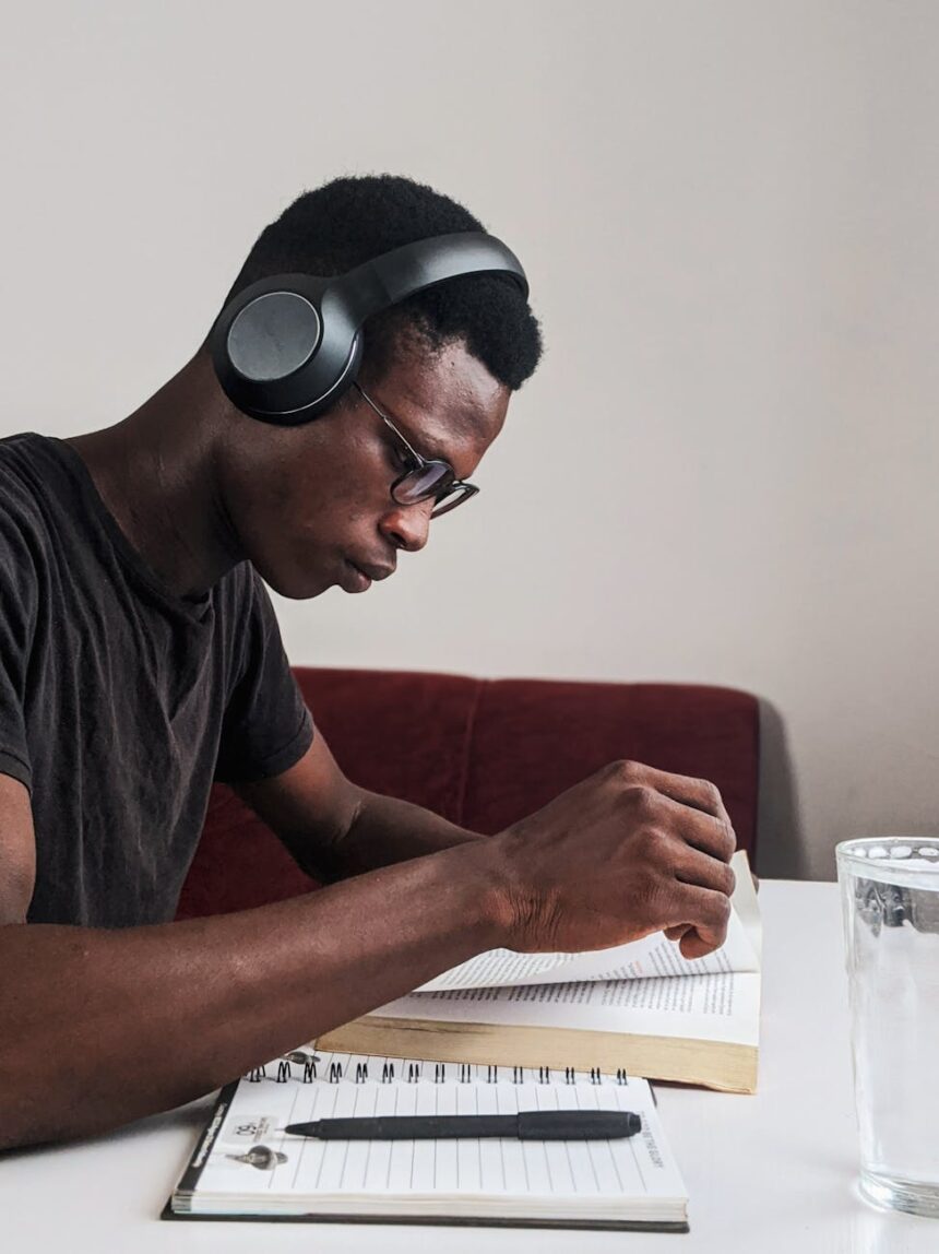 A focused student sits at a wooden desk, writing down academic goals in a spiral notebook surrounded by a stack of books.