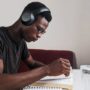 A focused student sits at a wooden desk, writing down academic goals in a spiral notebook surrounded by a stack of books.