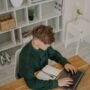 A focused student at a library desk using a laptop and tablet for digital note-taking.