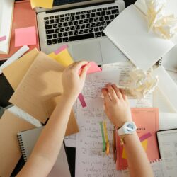 A stressed college student with their head in their hands, surrounded by textbooks and a laptop on a cluttered desk.