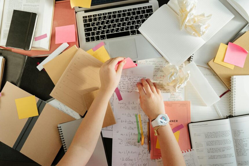 A stressed college student with their head in their hands, surrounded by textbooks and a laptop on a cluttered desk.