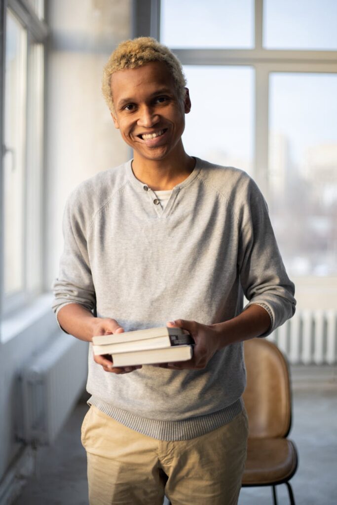 A frustrated student looks longingly out a window at a sunny day, a large pile of books on their desk.
