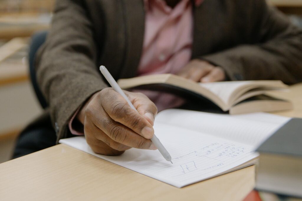 A focused student with headphones on, writing diligently in a notebook in a sunlit library.