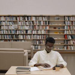 A focused student with headphones on, writing in a notebook at a library desk piled with books.