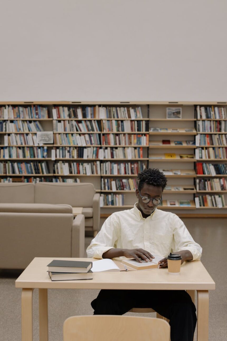 A focused student with headphones on, writing in a notebook at a library desk piled with books.