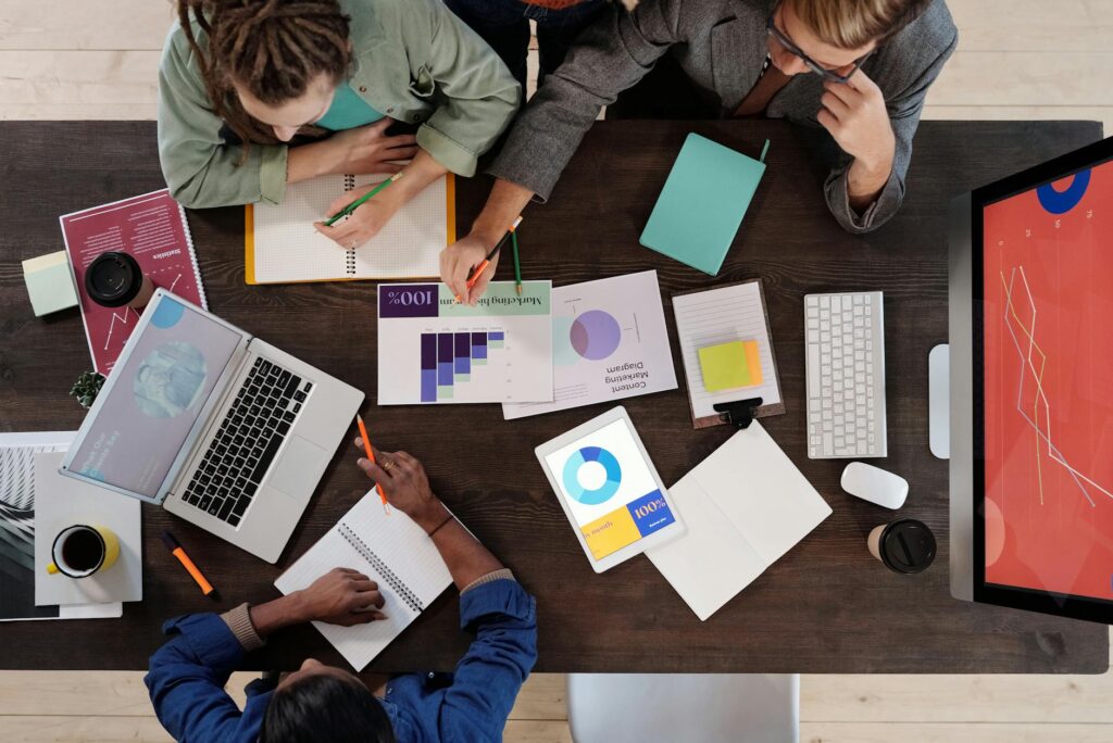 A diverse group of three students working together around a table, pointing at a laptop screen and smiling.