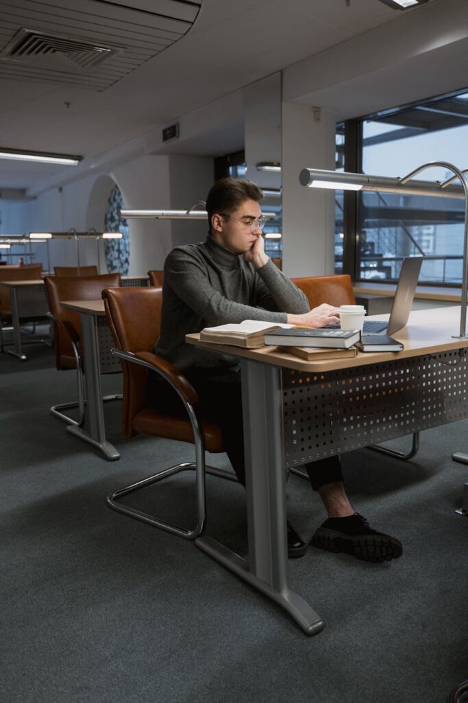 A young university student with a worried expression looking at their laptop screen next to a pile of bills.