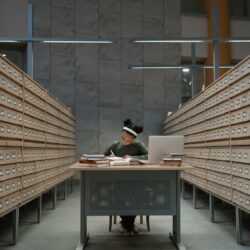 A focused university student sitting at a library desk, surrounded by stacks of books while researching grants.