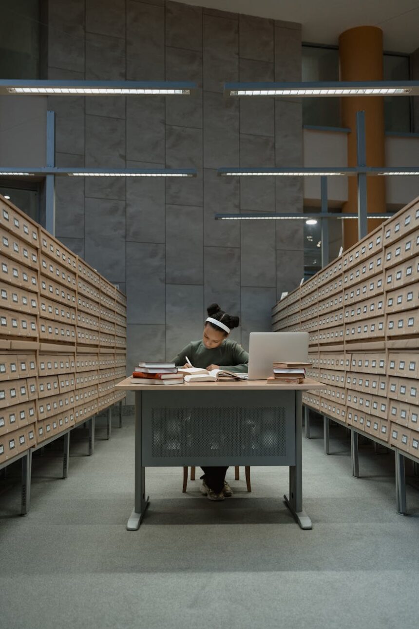 A focused university student sitting at a library desk, surrounded by stacks of books while researching grants.