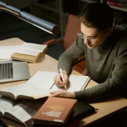A student sits at a desk with an open laptop, a tablet displaying organized notes, and a neat stack of physical notebooks.