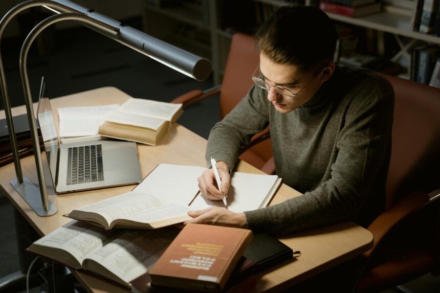 A student sits at a desk with an open laptop, a tablet displaying organized notes, and a neat stack of physical notebooks.