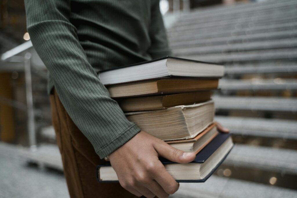 A tired-looking student with their head in their hands, surrounded by a large pile of books, showing the effects of poor time management.