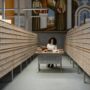 A focused university student with headphones on, writing in a notebook at a large library table.