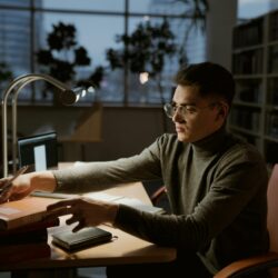 A university student diligently studying at a desk piled high with textbooks and notes, highlighting text.