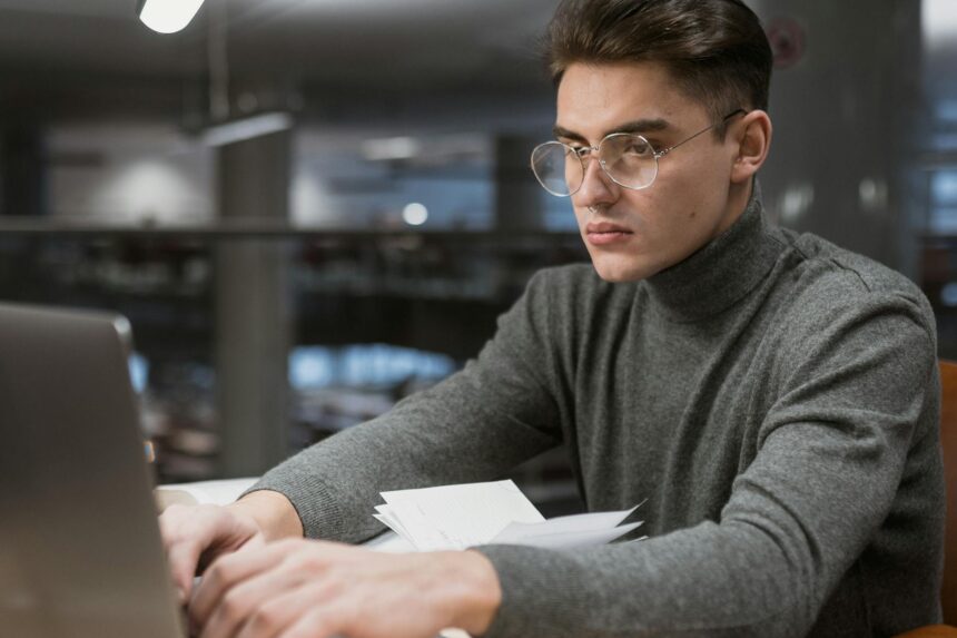 A focused college student sitting in a library, looking at investment information on their laptop.