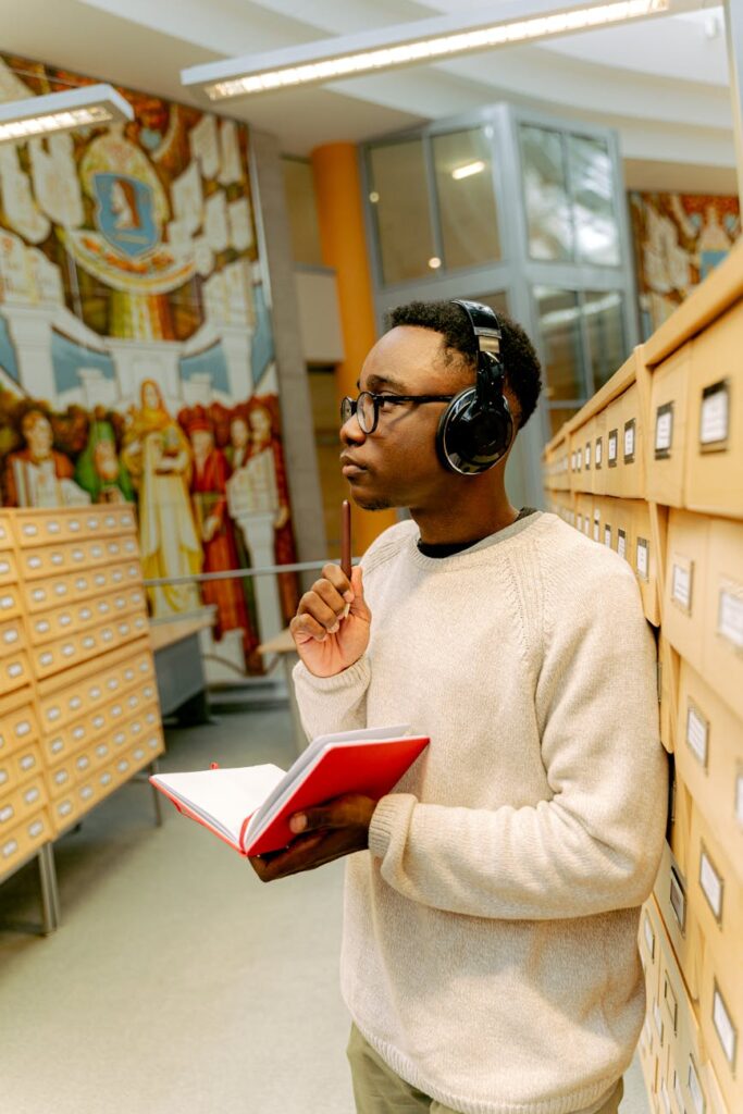 A focused young person sits at a library table, taking detailed notes from a book into a spiral notebook.