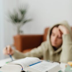 A focused student with headphones on, writing in a notebook at a sunlit desk.
