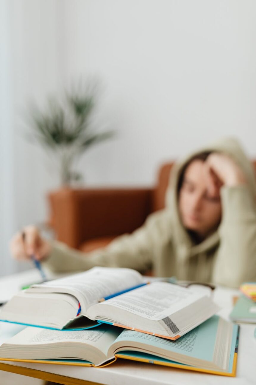 A focused student with headphones on, writing in a notebook at a sunlit desk.