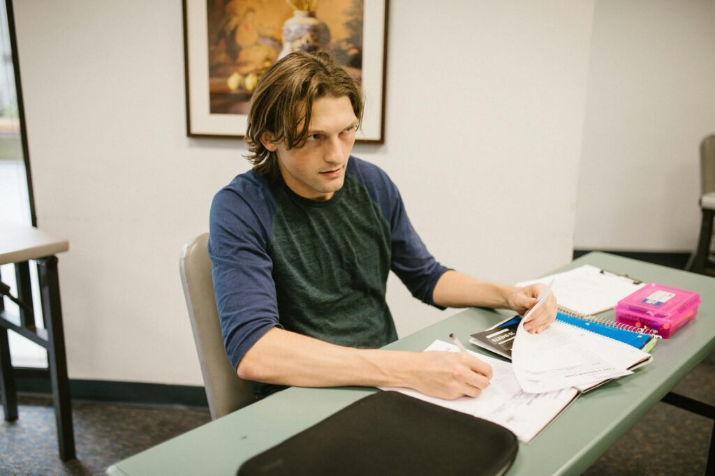 Close-up of a college student's hand writing neatly in a spiral notebook in a quiet university library setting.