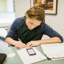 A stressed college student sits at a desk cluttered with textbooks and receipts, holding their head in their hands while looking at a laptop.
