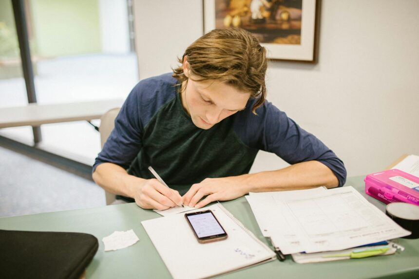 A stressed college student sits at a desk cluttered with textbooks and receipts, holding their head in their hands while looking at a laptop.