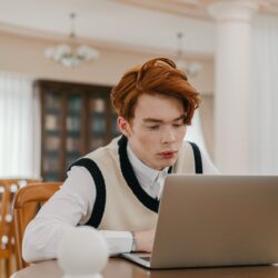 A university student at a cluttered desk, looking thoughtfully at their open laptop.