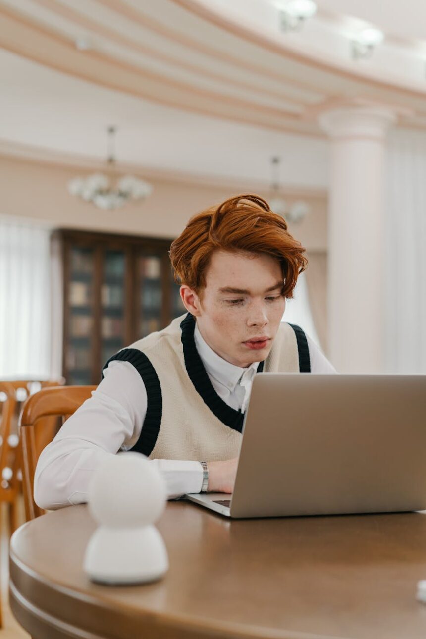 A university student at a cluttered desk, looking thoughtfully at their open laptop.