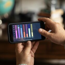 A young student sits at a desk, looking thoughtfully at an investment app on their smartphone.