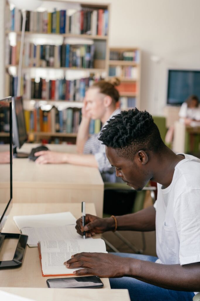 A young woman with glasses concentrating deeply as she writes notes in a spiral notebook in a quiet university library.