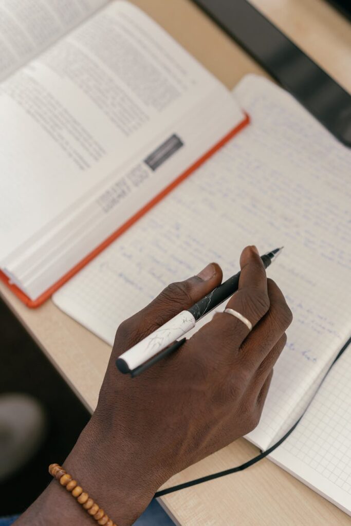 A focused young student sitting at a library desk, highlighted by a desk lamp, deeply engaged in studying for their exams.