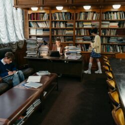 A focused student wearing headphones studies diligently at a desk covered in books and a glowing laptop screen.