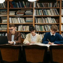 A focused college student sitting at their desk in a dorm room, working on a side hustle on their laptop.