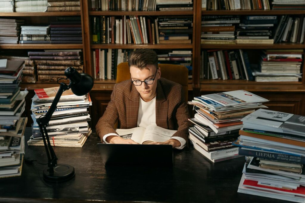 A student looking overwhelmed while sitting at a desk surrounded by a tall stack of diverse college textbooks.